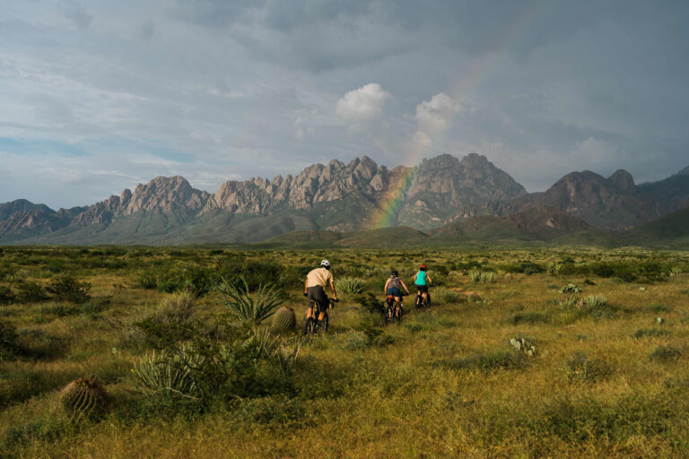 three mountain bikers riding a lush green trail with the jagged organ mountains in the distance and a rainbow