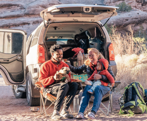 A multiracial family, mom, dad, and son, sitting on camp chairs behind their van enjoying the outdoors
