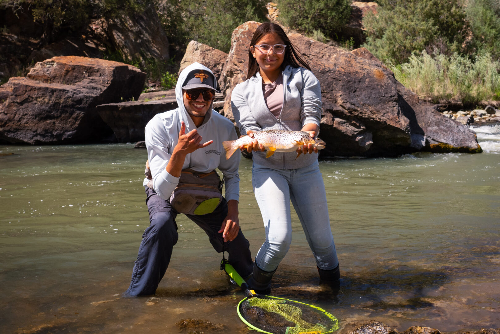 Young woman standing in river holding a large fish and young man standing next to her both looking excited