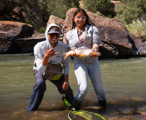 man and young woman standing in a river with the woman smiling holding a large fish