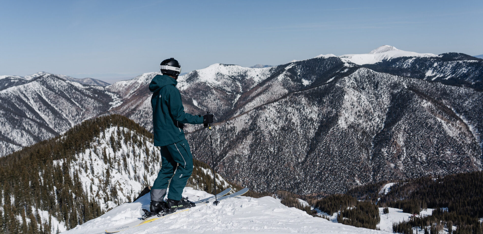 Person in blue snow clothes standing on a mound of snow with their skiis on overlooking a scenic vista of snow covered mountains