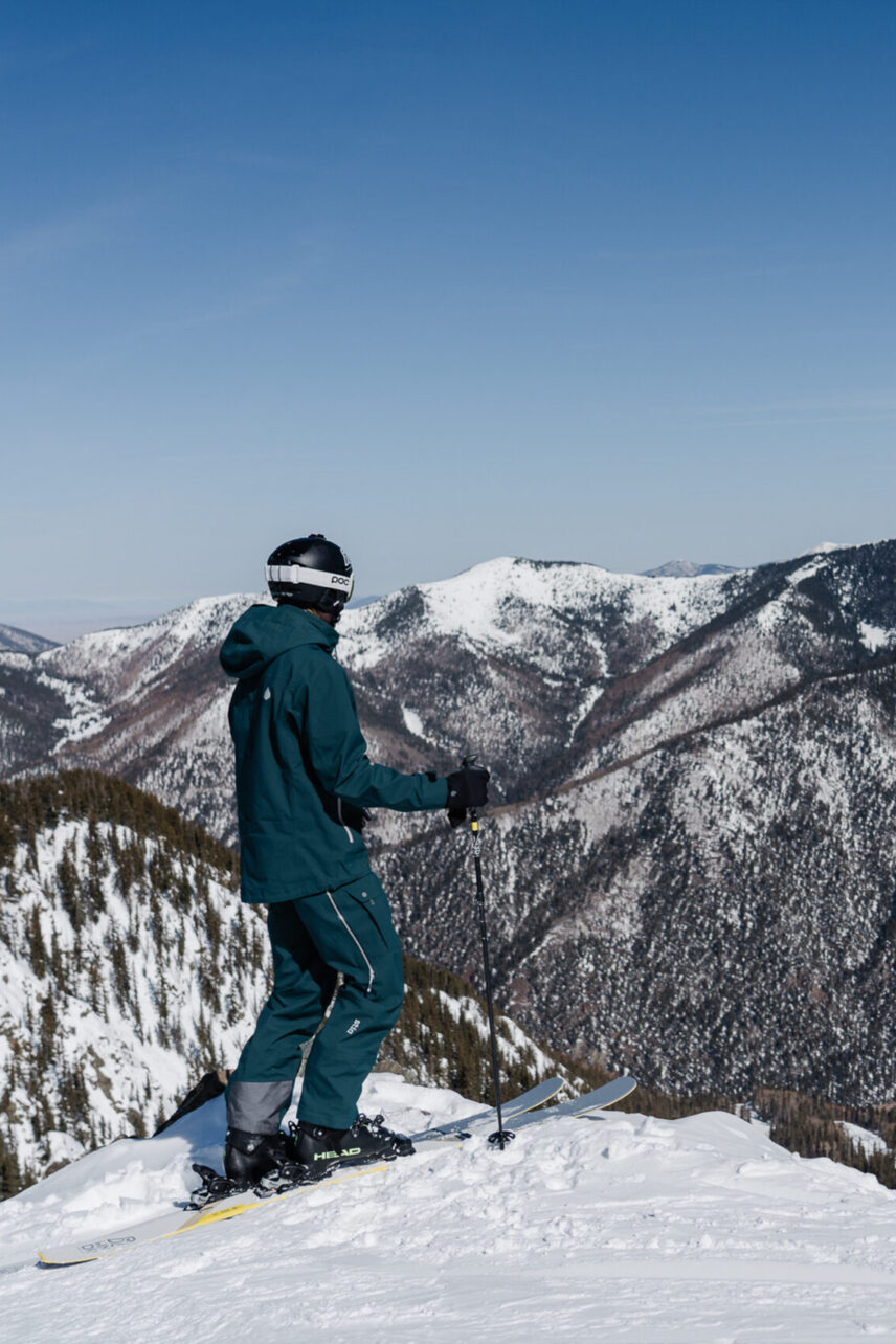 Mobile sized image of Person in blue snow clothes standing on a mound of snow with their skiis on overlooking a scenic vista of snow covered mountains