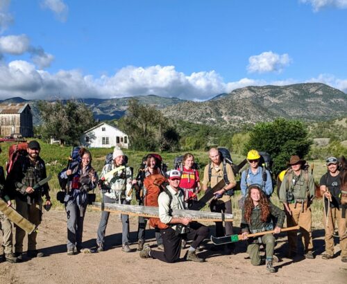 Large group of trail workers holding various tools and posing with a historic barn in the background