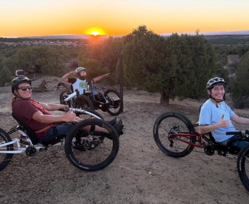 3 youth in recumbent adaptive mountain bikes smiling on the trail at sunset