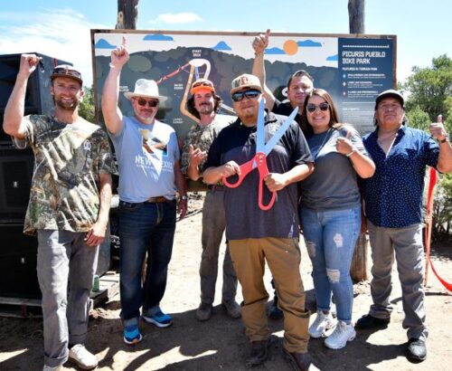 A group of people celebrating in front of a trail map sign with a man holding giant ribbon cutting scissors