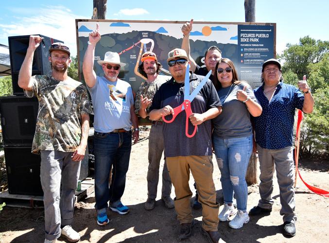 A group of people celebrating in front of a trail map sign with a man holding giant ribbon cutting scissors