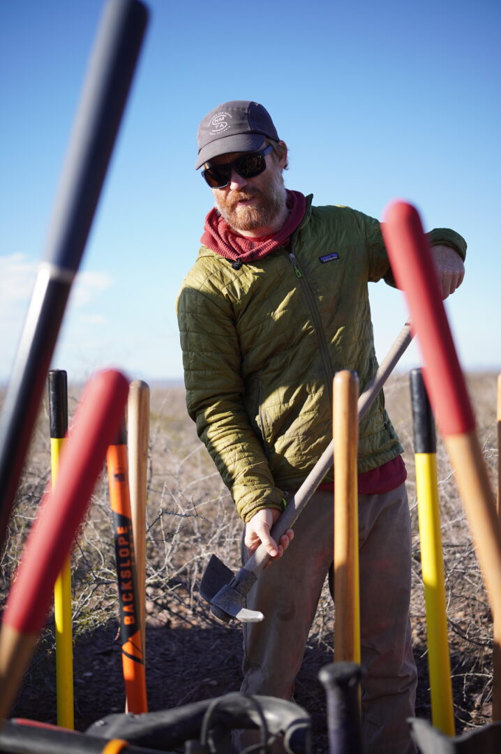 A man picking up a trail tool with handles from many other tools framing him in the photo