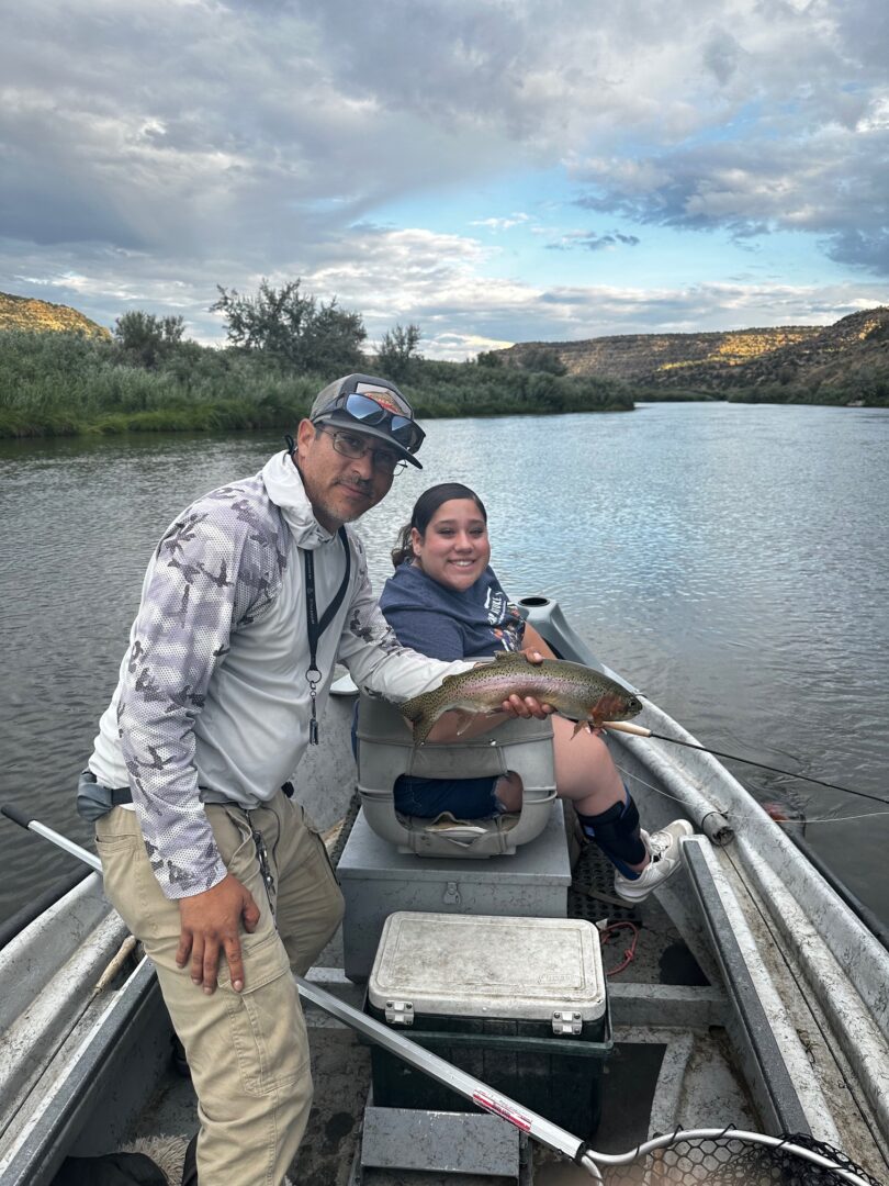 A man standing in a boat holding a trout with the girl who caught it sitting in the background smiling holding a fishing rod