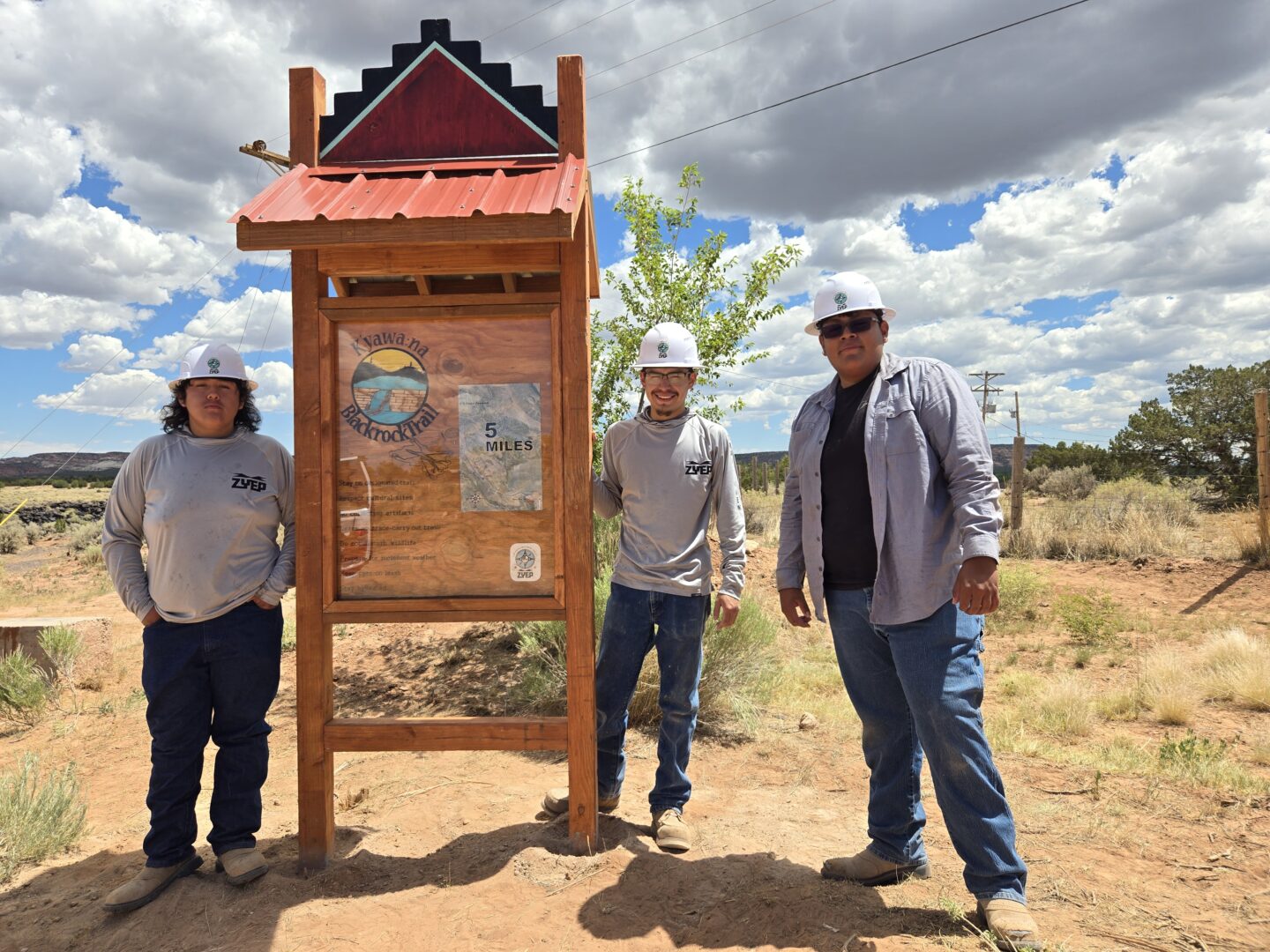 Three young people wearing hard hats standing proudly next to a wooden trail sign they just installed