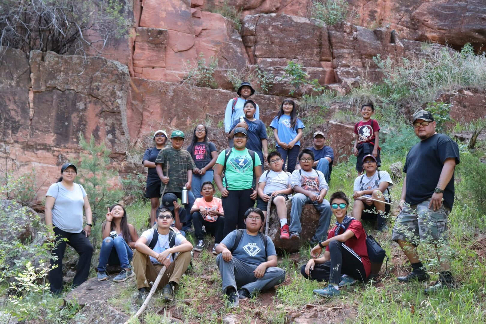A group of children pose in front of a red rock wall smiling and wearing backpacks
