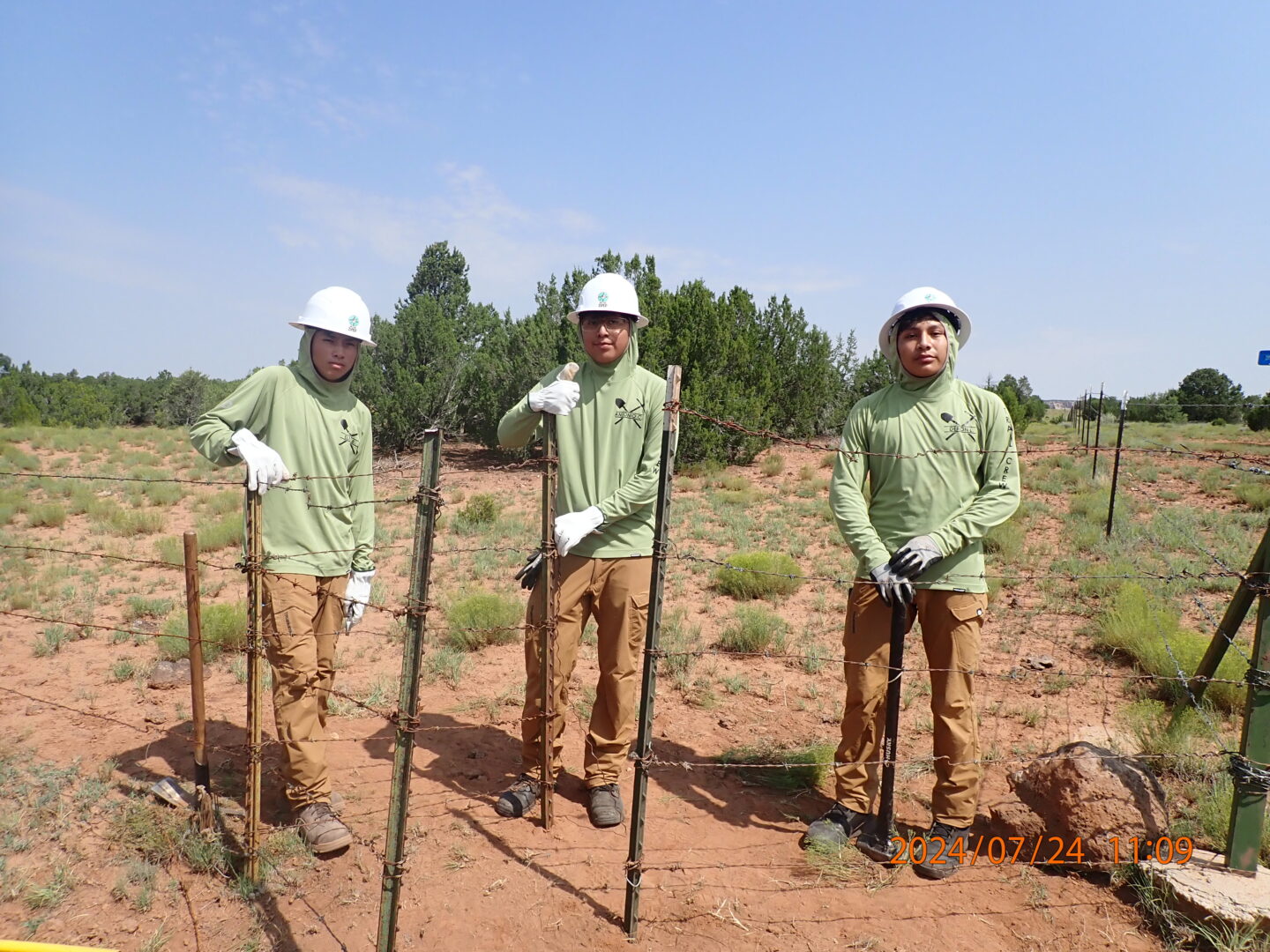 Three young men wearing green sun shirts, beige work pants, hard hats and gloves holding posts with barbed wire looking at the camera