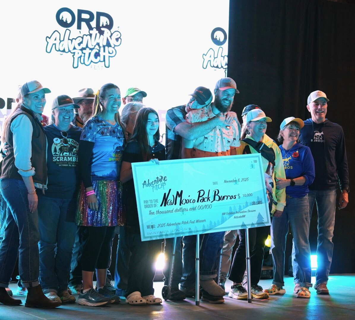 Man smiling holding a giant check on a stage surrounded by people smiling and embracing