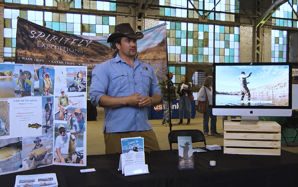 A man wearing a cowboy hat standing behind a booth showcasing his fly fishing business