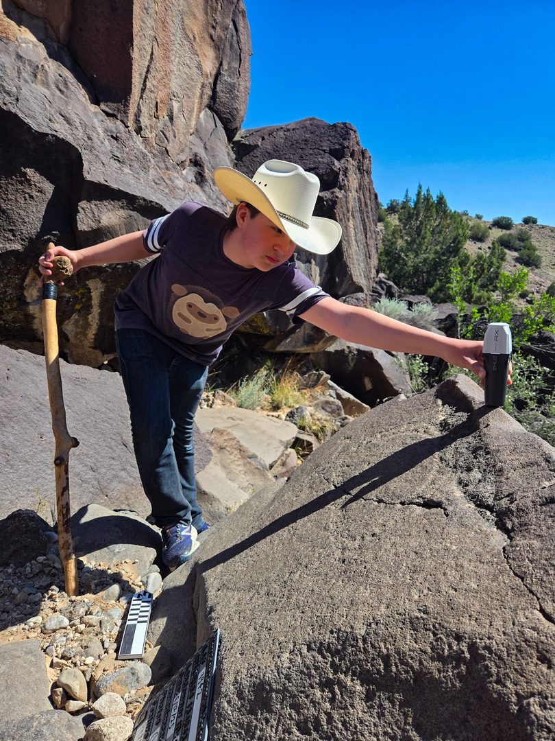 A young person wearing a cowboy hat leans over a petroglyph with a gps device