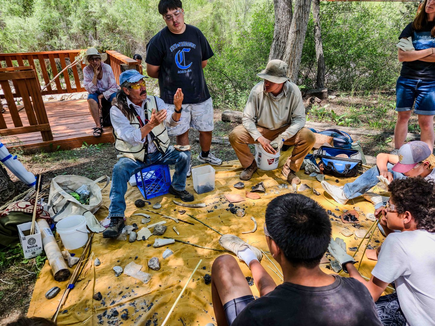 A man sitting on a milk crate surrounded by fragments of rock and bone doing a demonstration for a group of youth seated around him