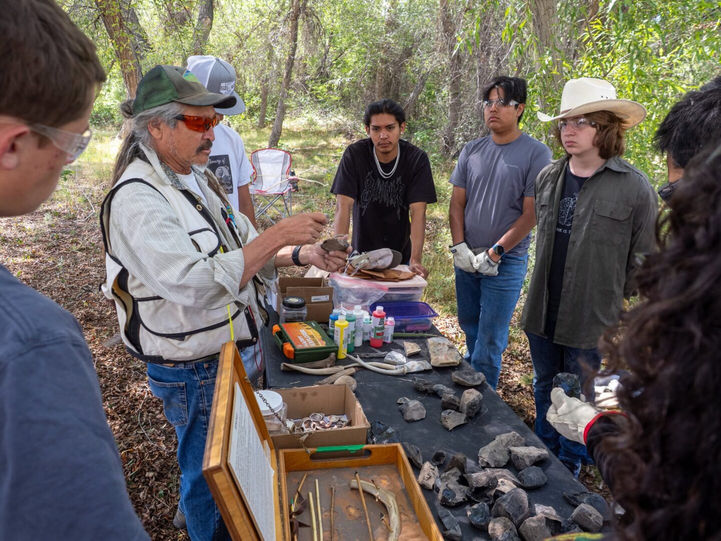 Teens standing around a table outdoors watching an elder demonstrate flint knapping