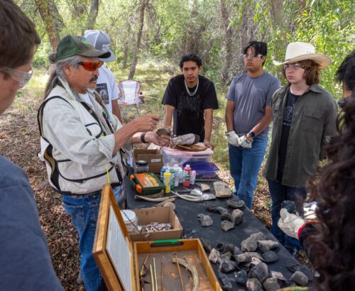 Teens standing around a table outdoors watching an elder demonstrate flint knapping