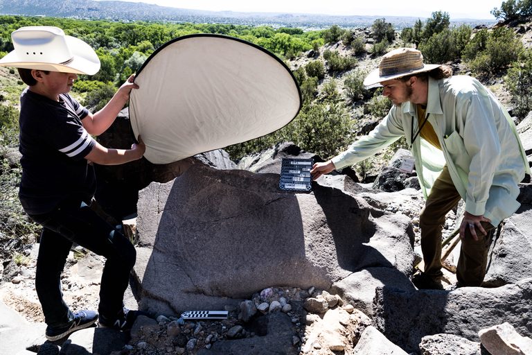 A young person shielding the sun with a reflector as another young person holds a tablet with notes on it over a petroglyph